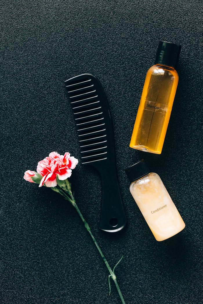 why-choose-us-img Flat lay of hair care essentials with a comb, bottles, and pink flower on dark background.